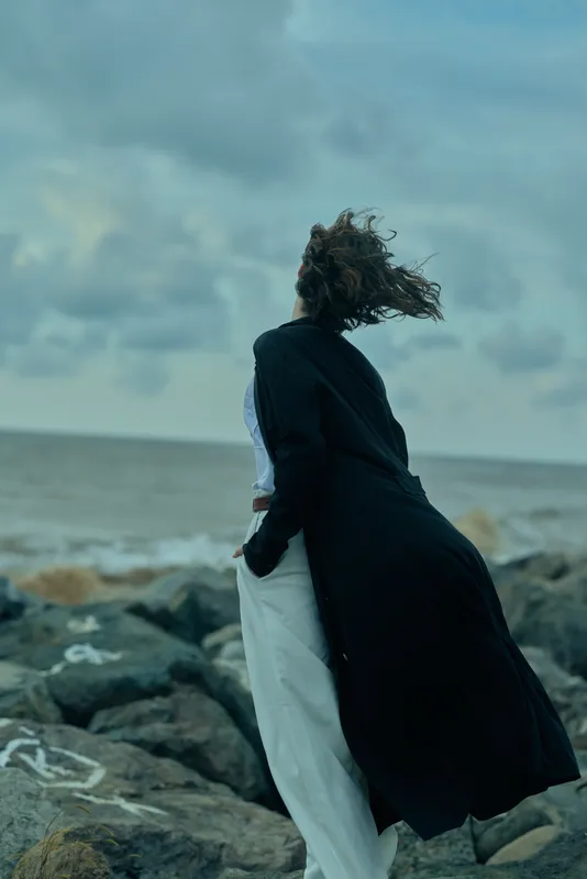 Windy coastal scene with a figure in a long black coat standing on jagged rocks by the sea, hair blown back, white pants visible, a moody sky and sense of movement, solitude and dramatic ocean