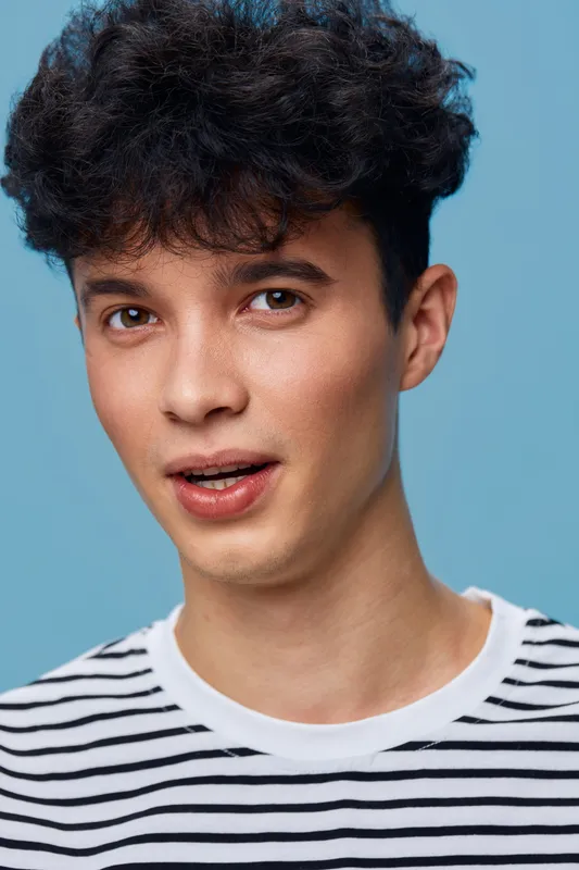 young man with curly hair wearing striped shirt against blue background, expressing subtle surprise or curiosity, casual style portrait with soft lighting