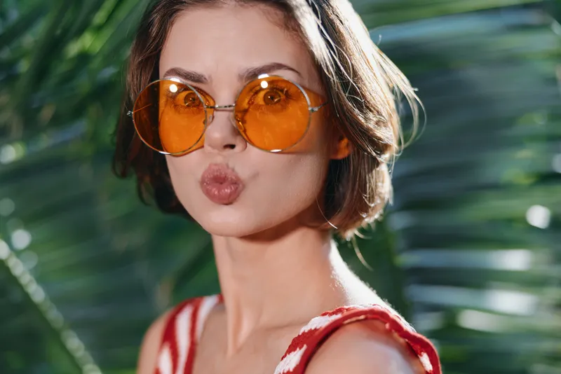 A stylish woman wearing orange sunglasses puckers her lips playfully in a sunny outdoor setting, capturing a vibrant, carefree mood with tropical greenery in the background.