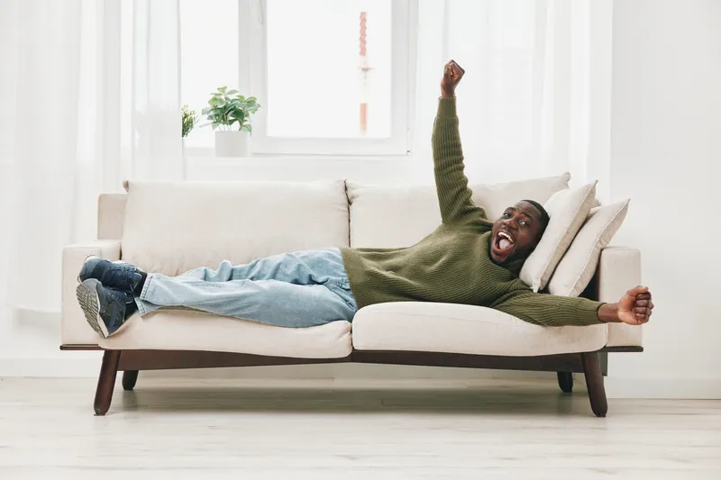 Happy Black man relaxing on a cozy sofa, expressing joy and comfort in a bright, modern living room with soft natural light