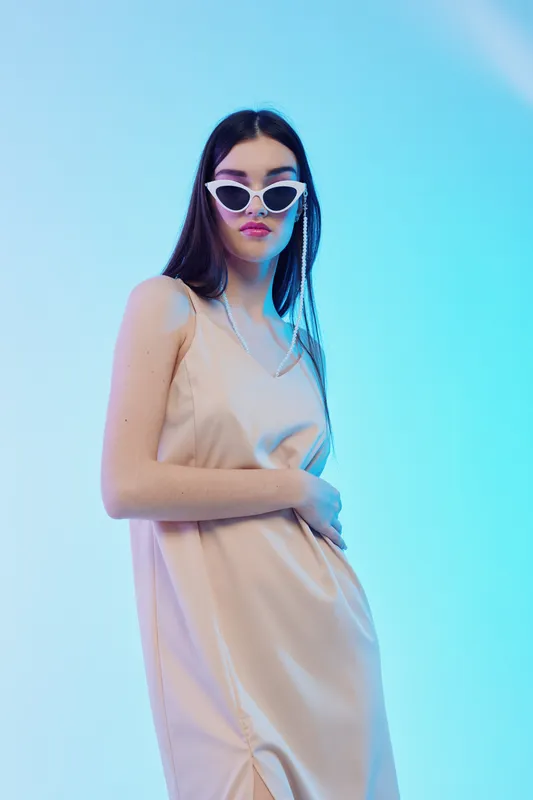 Young woman in a beige dress and white sunglasses poses confidently against a blue gradient background. Concept of woman beauty fashion and modern style.
