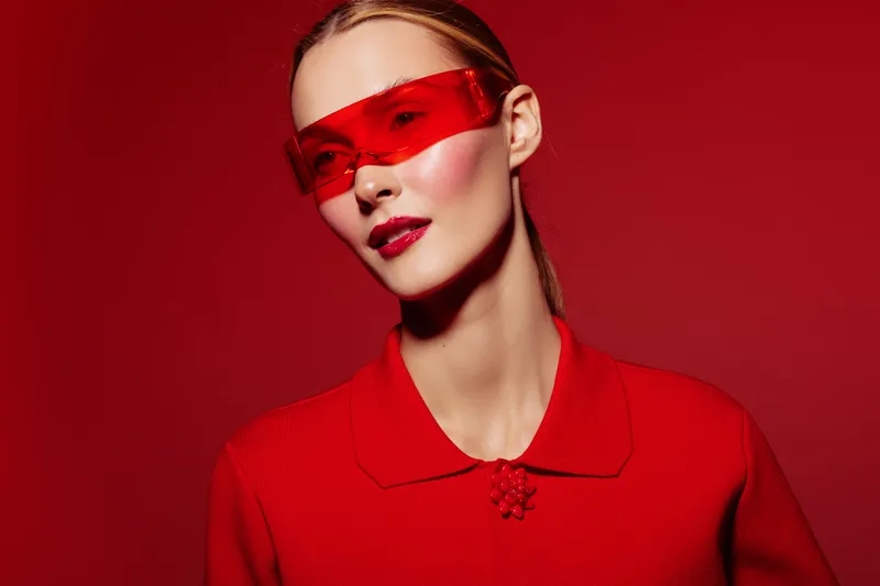 Stylish woman in red sweater and futuristic red glasses poses against red background. Fashion portrait with bold makeup and sleek hairstyle. Concept of modern style and confidence.