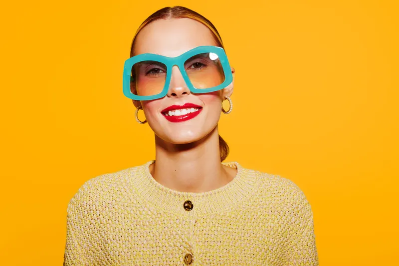 Smiling woman with large blue sunglasses and red lipstick wears yellow knitted sweater against bright yellow background. Concept of fashion, style, beauty, and vibrant colors.