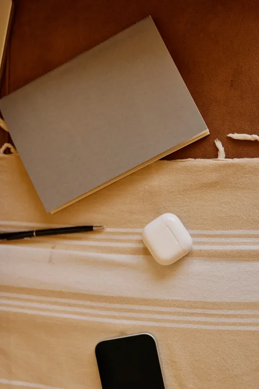 Cozy home workspace, flat lay with notebook, pen, phone and wireless earbud case on a striped fabric surface, warm lighting, calm atmosphere, simple organized setup