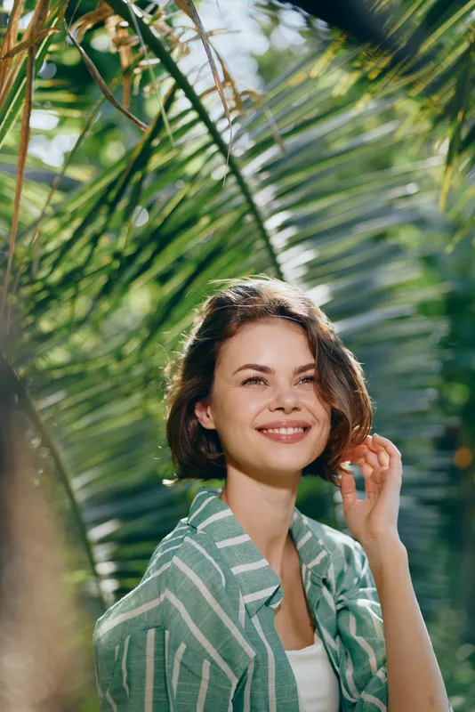 Portrait of a smiling woman outdoors among tropical palm leaves, bathed in sunlit light, wearing a casual striped shirt.