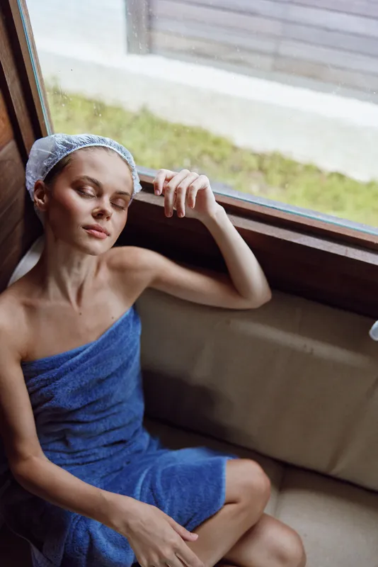 relaxing woman wrapped in blue towel with shower cap resting by window, enjoying serene moment indoors with natural light and soft ambiance.