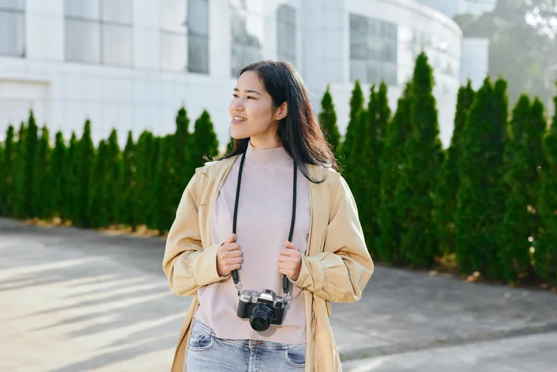 Smiling woman in a light coat outdoors with a camera hanging from her neck, casual street style, exploring a bright campus setting, candid photography and travel vibe in sunny afternoon light