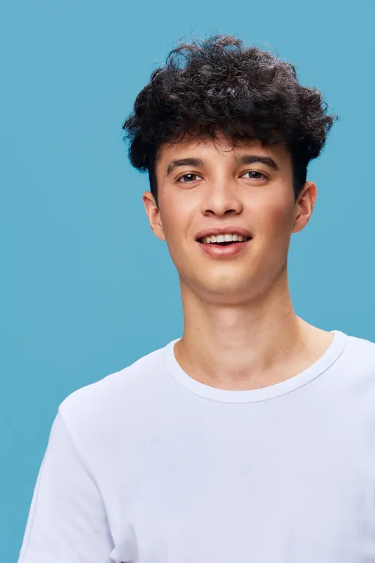 Young man with curly hair wearing a white t-shirt smiling confidently against a bright blue background, radiating positive energy and youthful charm.