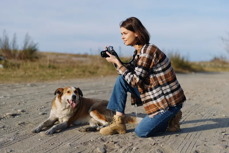 Woman kneels with camera in hand, photographing a relaxed dog on a sunny dirt road, casual plaid jacket, boots, open landscape and calm outdoor moment.