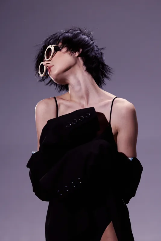 Stylish woman with short hair wearing oversized sunglasses and black clothing posing against purple background