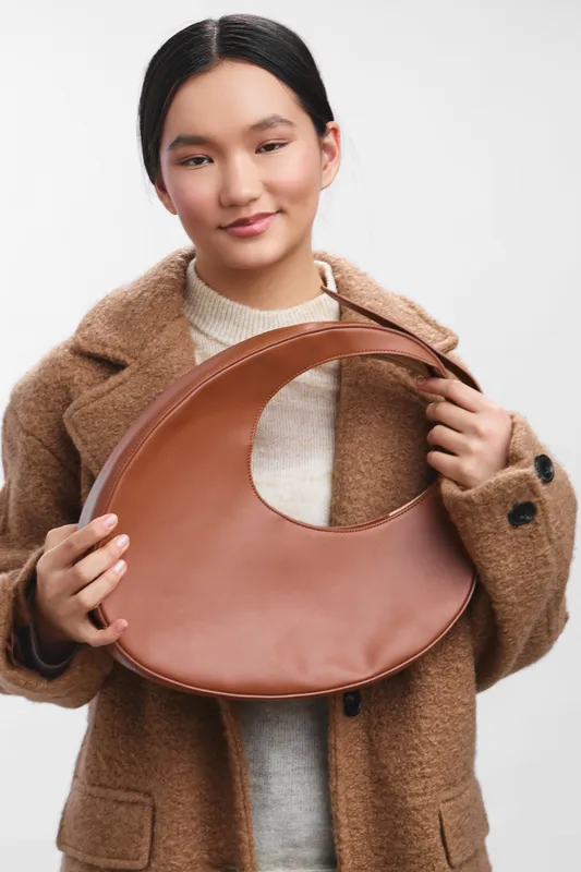 asian woman wearing a warm coat holds a chic brown leather bag in a clean studio setting, soft lighting highlighting texture and style. Her calm expression conveys confidence and elegance, suitable