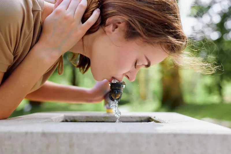 Woman drinking water from a public fountain in a park, young female leaning to sip fresh outdoor water on a sunny summer day, closeup portrait with blurred green background.