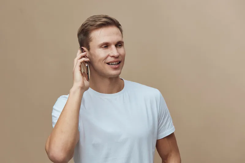 smiling caucasian man talking on smartphone holding hand near ear casual white t-shirt beige background communication technology concept