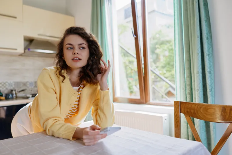 A young woman with curly hair sits at a sunlit kitchen table in a cozy home, looking thoughtful while holding a smartphone, ready to chat, plan the day and enjoy a calm moment.