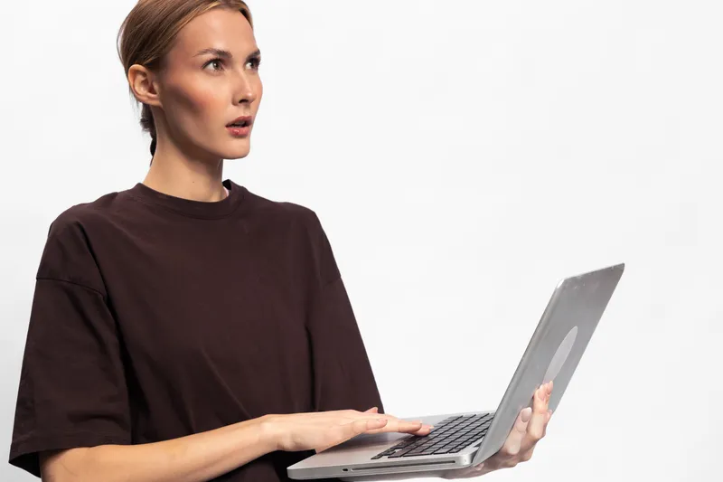 Surprised young woman in dark shirt holding laptop with one hand looks away. Isolated on white background. Concept of unexpected news, technology, and online communication.