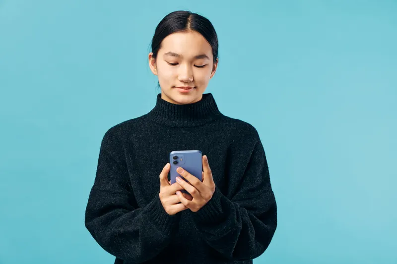Young woman in black sweater holding smartphone and looking at screen with slight smile on blue background in studio, technology, communication and casual style concept.