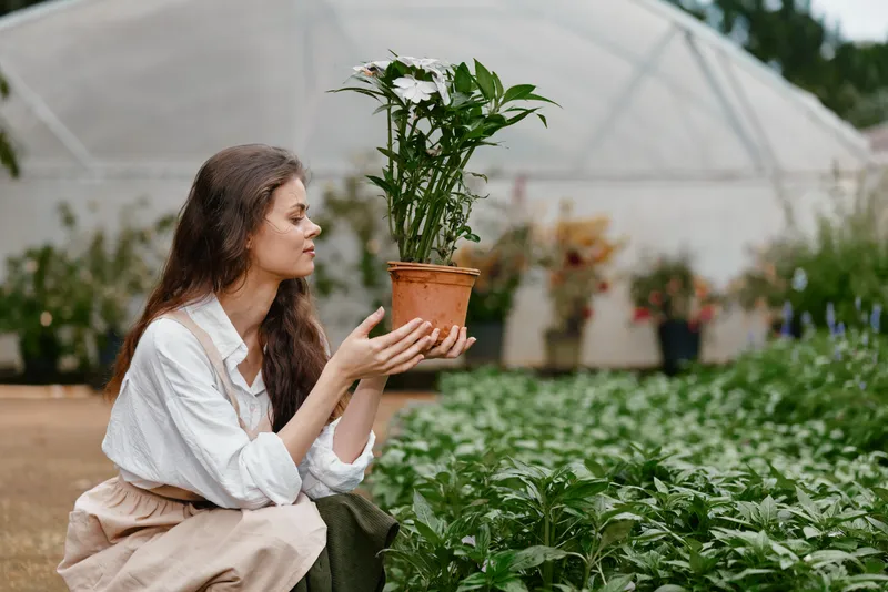 Young woman with a potted plant in a greenhouse, showcasing emotions of joy and tranquility amidst lush greenery in a warm, natural setting