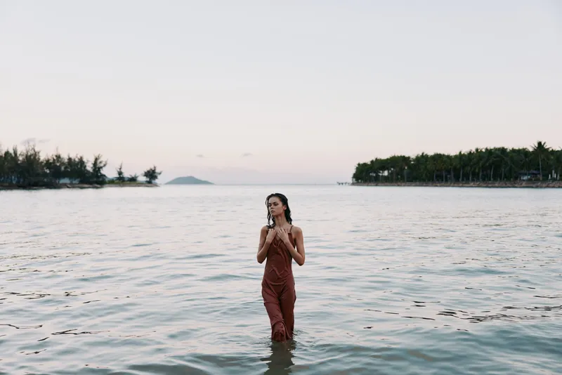 A lone figure stands waist-deep in calm coastal water at dusk, wearing earthy overalls, exuding calm, freedom, and a contemplative mood against a tranquil seascape.