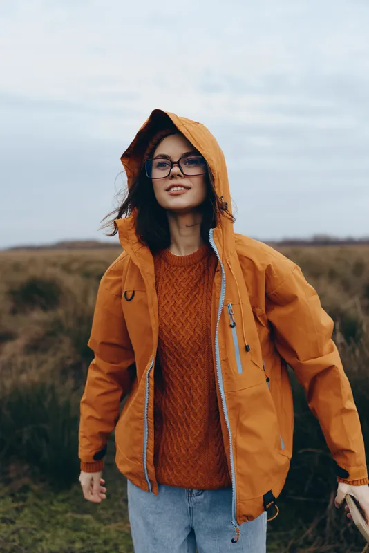 Woman lifestyle outdoors portrait hood orange jacket backpack hiking nature, smiling woman in orange raincoat with knit sweater walks on countryside trail under cloudy sky, travel concept