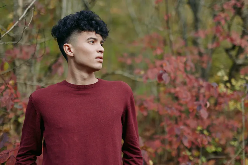 A candid portrait of a young man in a burgundy sweater among autumn foliage, showcasing authentic presence and credible expression suitable for editorial stock use.