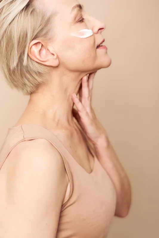 Mature woman with short blonde hair applies white face cream on cheek, touching neck gently. Concept of skincare, aging care, natural beauty, and wellness in soft studio lighting.
