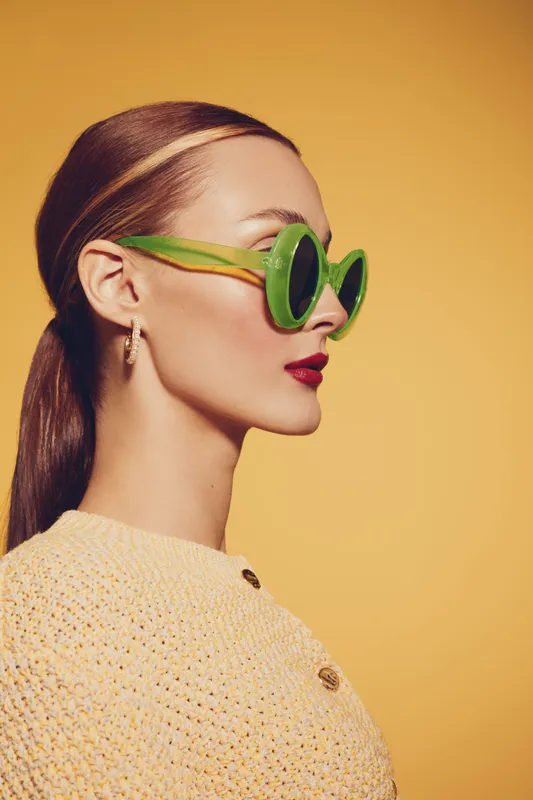 Stylish woman with green sunglasses and red lipstick poses in profile against yellow background. Fashion concept with retro accessories and elegant hairstyle in studio lighting.