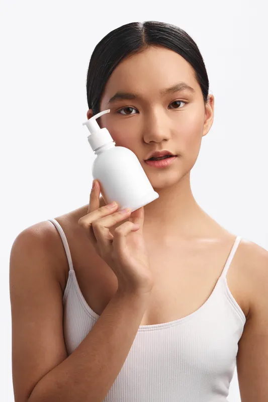 Asian woman holds a pump bottle against a clean white backdrop. She looks calm and focused, presenting a minimalist skincare or personal care product shot with a bright, studio feel.