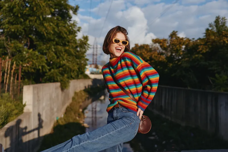 young woman in a striped sweater and sunglasses, smiling woman wearing denim jeans leans on concrete railing in urban outdoor setting, joyful expression, colorful knitwear and casual outfit.