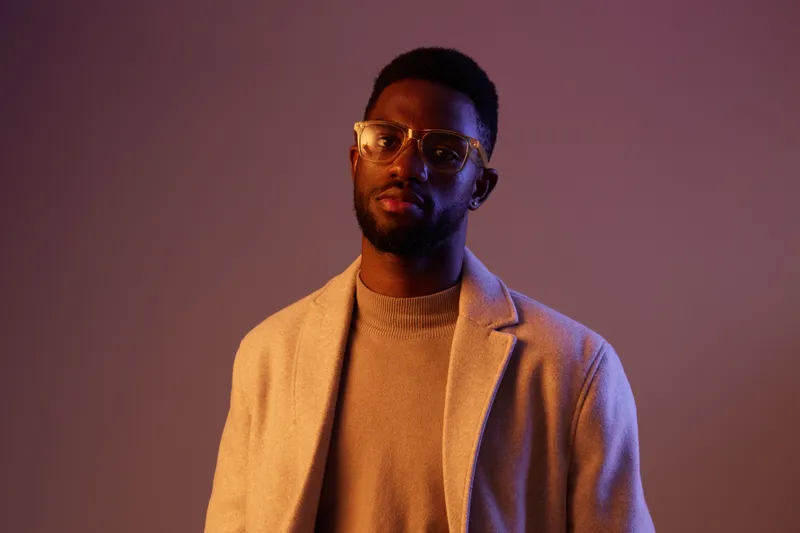 Stylish portrait of a Black male model wearing modern eyewear and a light sweater against a softly lit, gradient background, showcasing confidence and sophistication