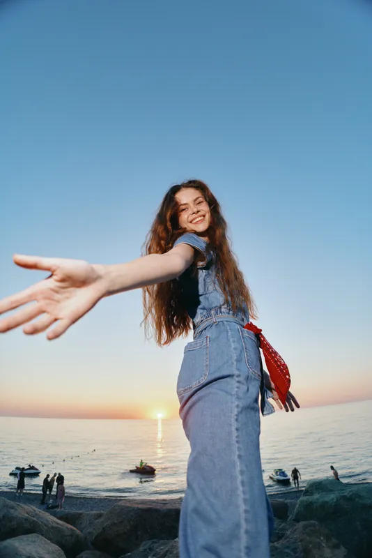 smiling woman in denim jumpsuit enjoying sunset by the sea with outstretched hand and relaxed mood during summer evening