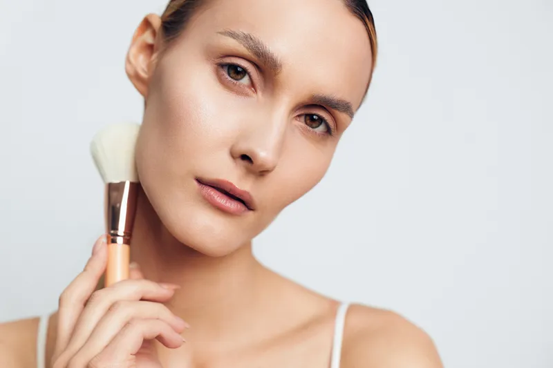 Woman applies makeup with brush on her cheek, beauty skincare closeup portrait in studio lighting, calm grooming and hygiene concept on clean pale background