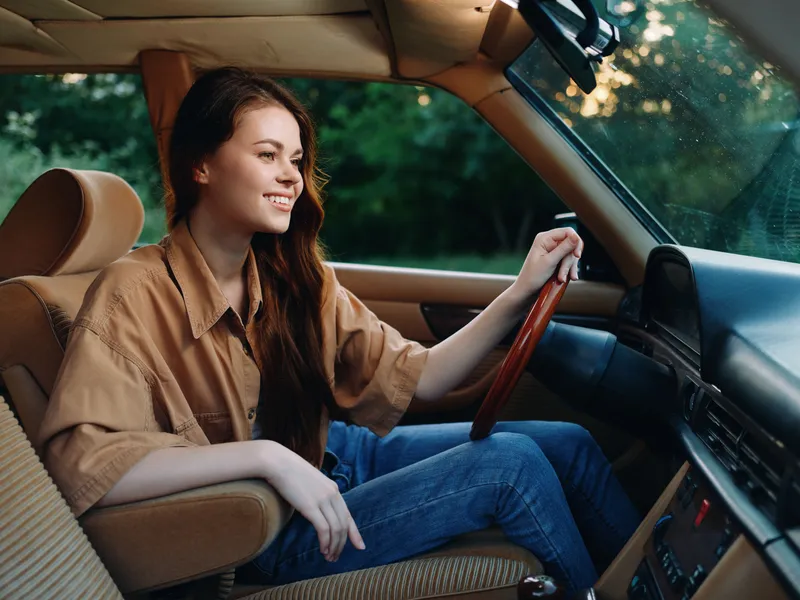 Young woman smiling while driving a classic car, wearing a casual beige shirt and denim jeans, showcasing joy and freedom in a green outdoor setting.