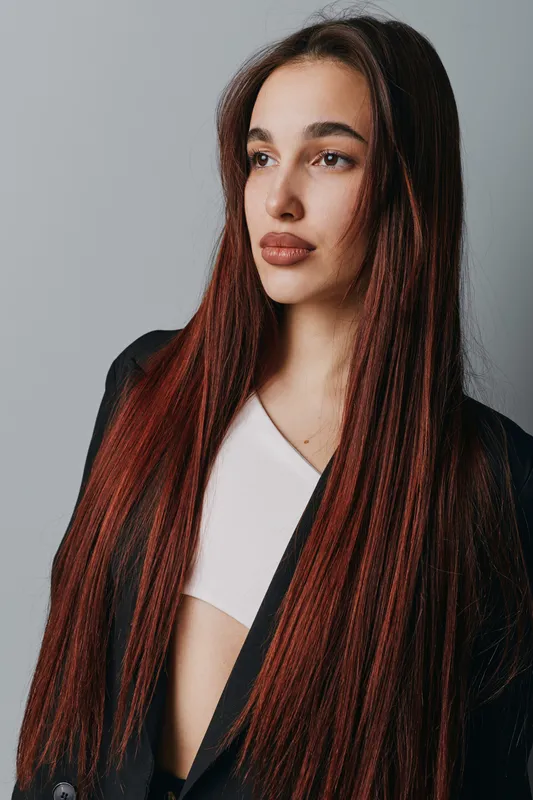 Young woman with long straight hair wearing black jacket and white top posing against gray background looking sideways with calm expression in studio portrait photography.