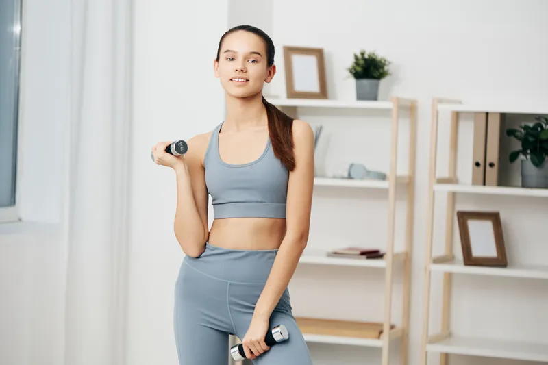 Young woman fitness athlete in gray sportswear lifting light dumbbells in a bright home workout setting with wooden shelving, plant, framed art, confident posture and healthy focus