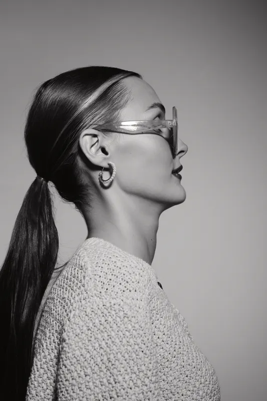Profile of a young Caucasian woman with pale skin and long dark hair wearing transparent glasses and a knitted sweater in studio.