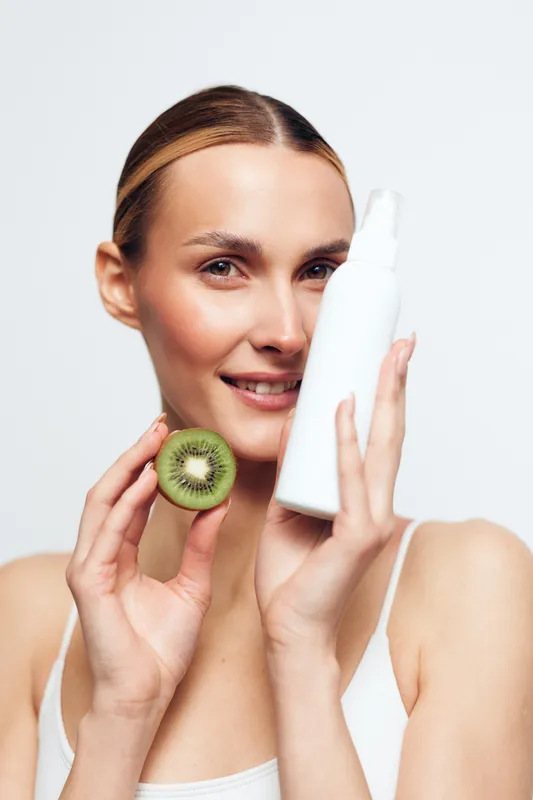 Young woman with blonde hair holds a white cosmetic bottle and a sliced kiwi near her face while smiling gently against a plain background.