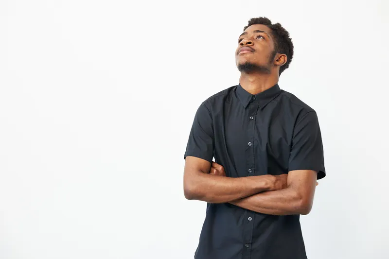 A thoughtful black man looking up while standing confidently with arms crossed, showcasing a blend of strength and introspection against a clean, bright background.