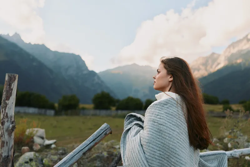 Cheerful woman outdoors, wrapped in a cozy blanket, gazing toward distant mountains, sunlit breeze, relaxed posture, natural colors, serene lifestyle moment