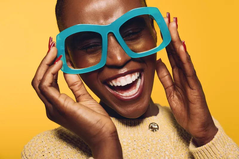 Happy African woman with dark skin wearing oversized blue glasses and yellow sweater smiles widely against yellow background. Concept of joy, fashion, and vibrant lifestyle.