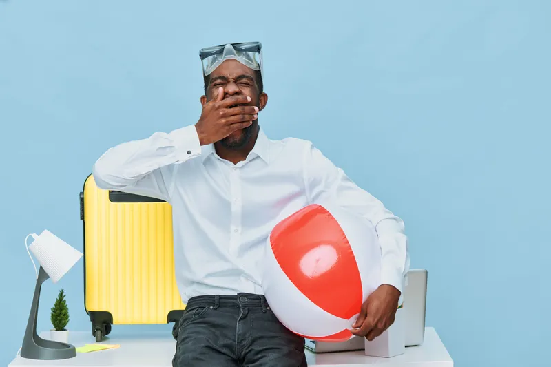 Casual smiling man with dark skin in white shirt holding a beach ball in a bright studio setting with travel accessories like a yellow suitcase and lamp, concept of vacation and relaxation.