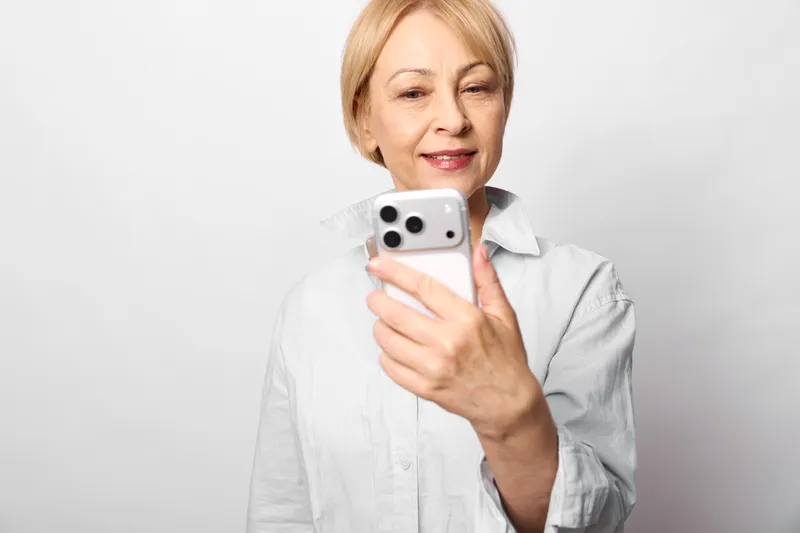 Woman senior smiling holds smartphone for selfie, camera portrait indoors on neutral white background, mobile communication and technology for everyday photo taking.