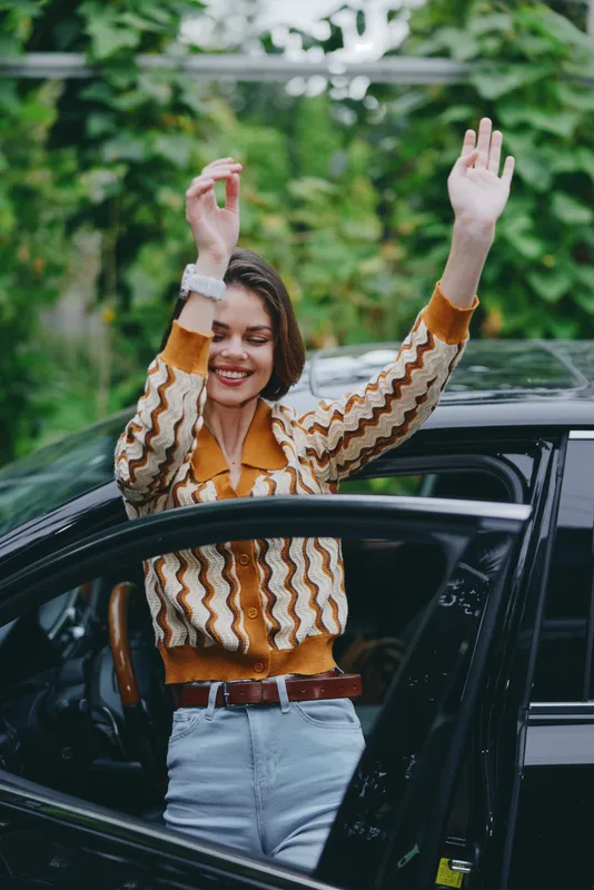 Joyful woman steps out of a car with arms raised in celebration, wearing a striped knit sweater and jeans; candid, energetic moment outdoors.
