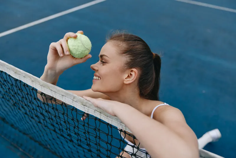 Young athletic woman smiling while holding a tennis ball on a blue court, showcasing energy and enthusiasm for sports and healthy living