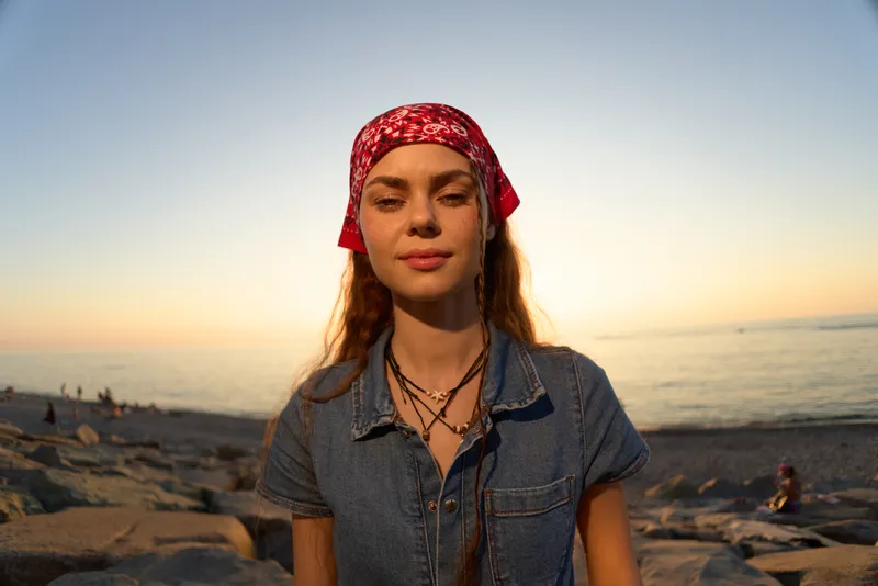Lifestyle woman wearing modern boho-western denim street style with a red bandana, standing outdoors near the beach at sunset with warm film color lighting.