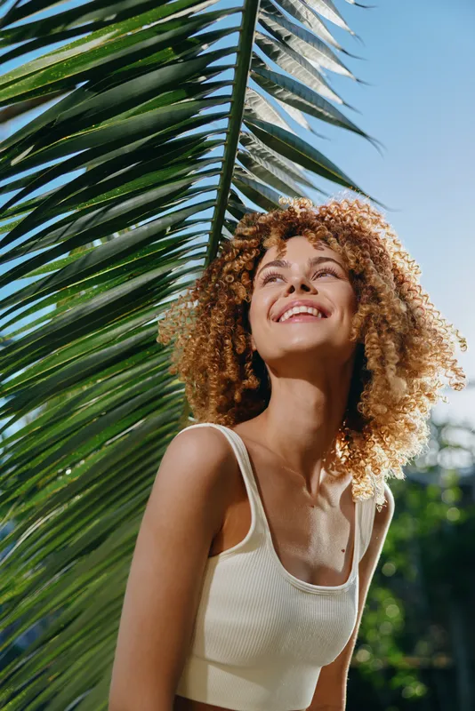 smiling woman with curly hair standing under palm leaves in bright sunlight outdoors, wearing a white tank top with blue sky background