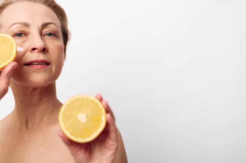 Mature woman holds two halves of a fresh orange close to her face. Clear skin and natural expression are emphasized against a white background. Concept of health and vitality.