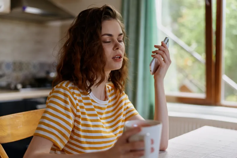 Young woman with curly hair sits at a sunlit kitchen table in a bright home, wearing a yellow striped tee, looking at her phone with a calm, thoughtful smile, a mug nearby.