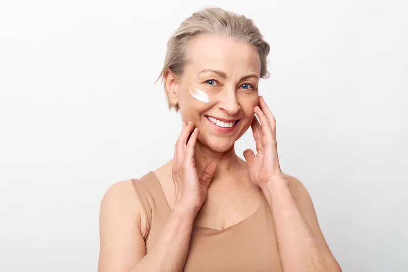 Smiling middle aged woman with short blonde hair applies white face cream on cheek. Studio portrait on white background. Concept of skincare, beauty, wellness, and natural self care.