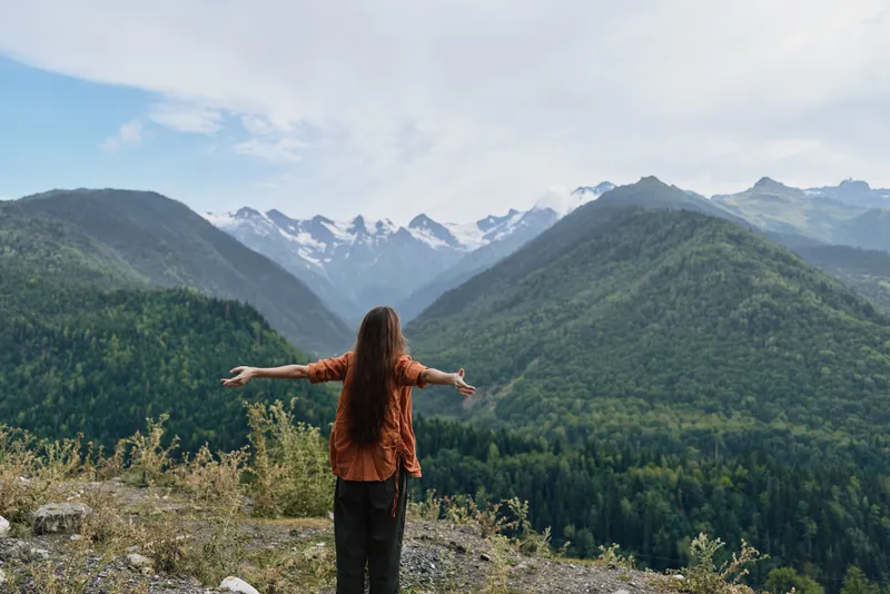 Person stands on a rocky overlook with arms outstretched, embracing a vast mountain landscape and lush forest below, conveying freedom, adventure, and a peaceful connection with nature.