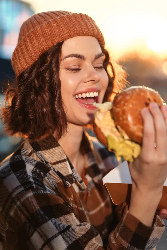 Woman burger smile outdoors beanie plaid enjoying a juicy burger in warm golden hour glow, candid lifestyle portrait with authenticity and emotional storytelling, cozy urban mindful living moment.